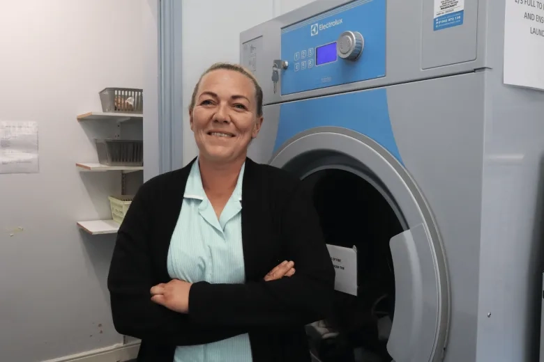 Picture of a housekeeper smiling next to a washing machine  with her arms crossed