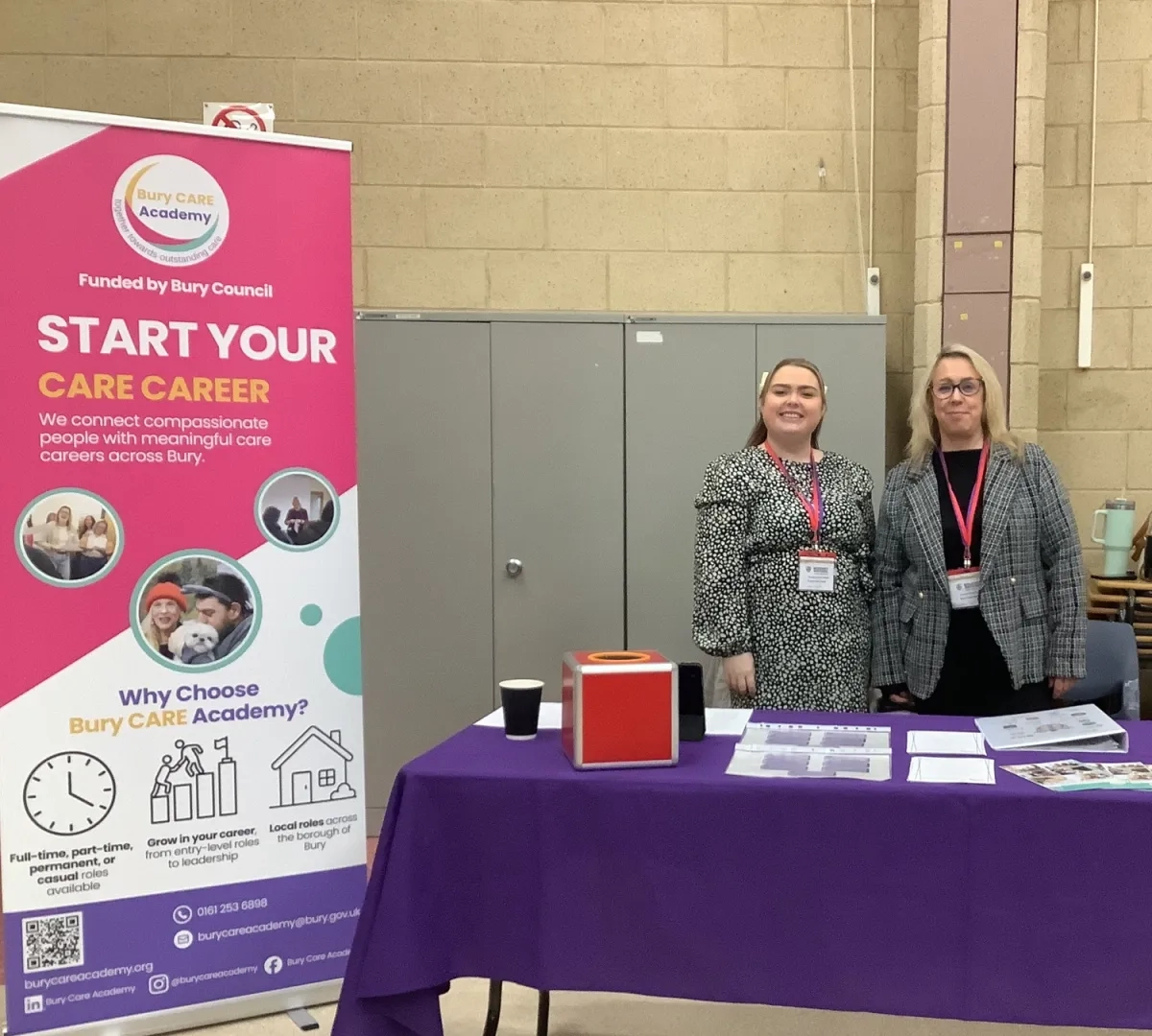 Two Bury Care Academy staff members pictured next to a Bury Care banner at a Woodhey highschool event