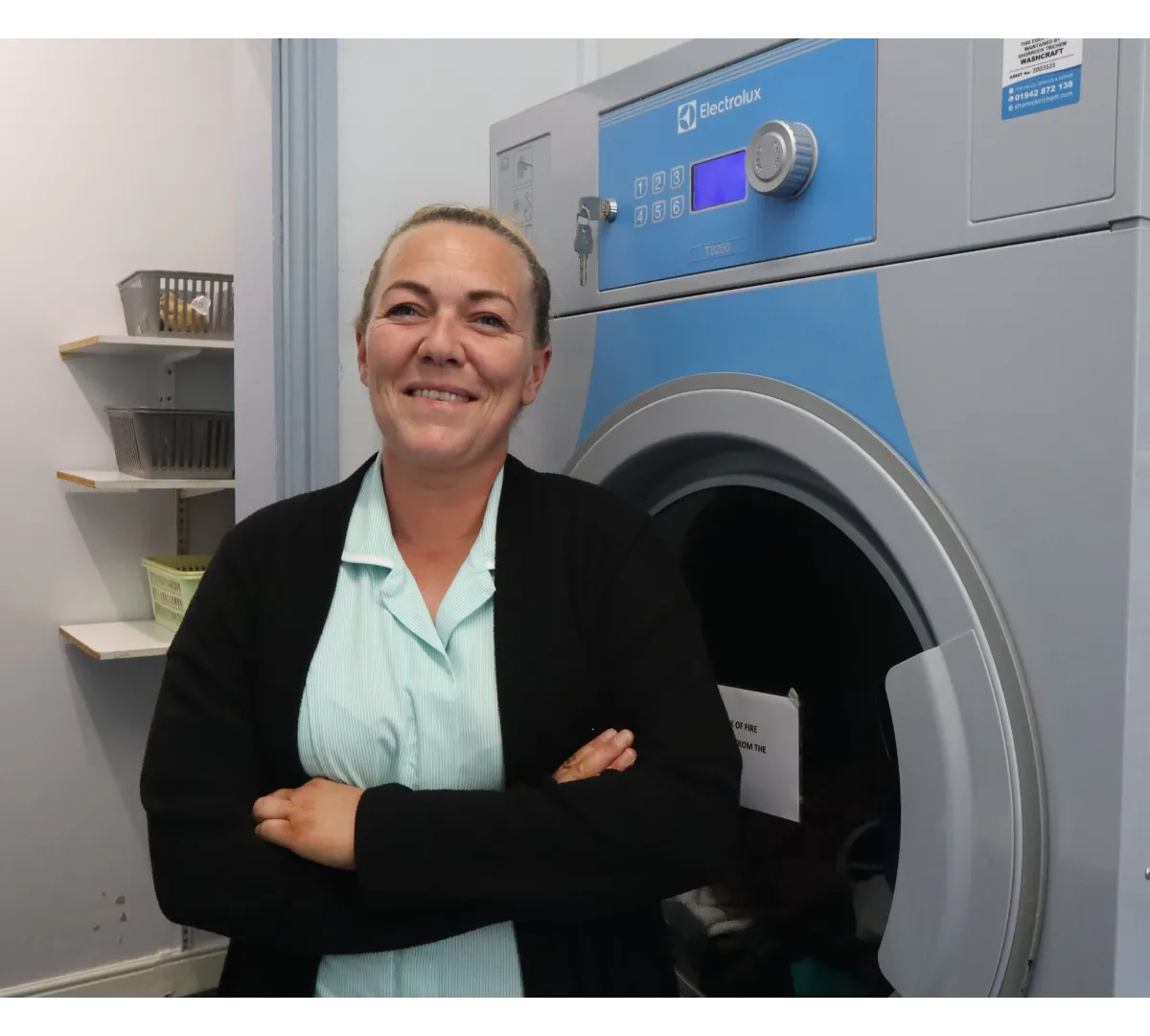 Care home housekeeper smiling next to a washing machine, supporting the cleanliness and comfort of residents.