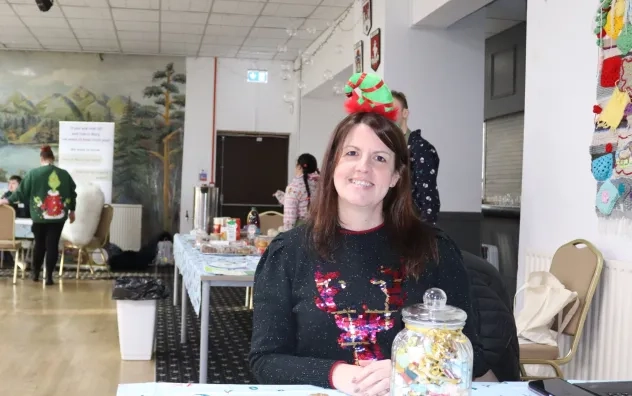 woman at the front desk of the persona Christmas fair smiling and wearing Christmas decorations, guests can be seen in the background preparing for event