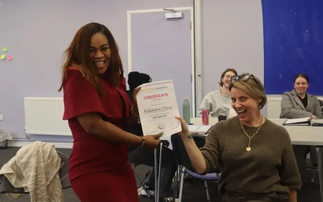 Image shows participant on the LEAD Programme smiling getting their certificate. Lead trainer on the programme in pictured smiling while presenting the certificate on one knee.