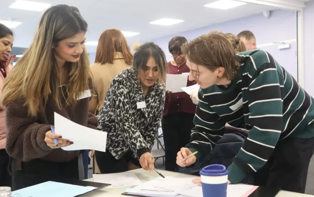 lead programme participants working on a task together, the girl stood in the middle points towards the table while the other two participants look and smile