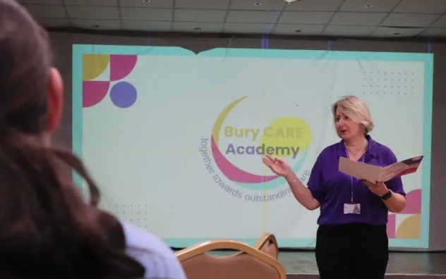 presenter speaking to attendees at at event, bury care academy logo is shown on the screen behind the presenter