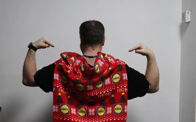 A man at the Christmas Fair with his back turned to the camera facing the camera is a Lidle Christmas jumper being shown draped over his back.
