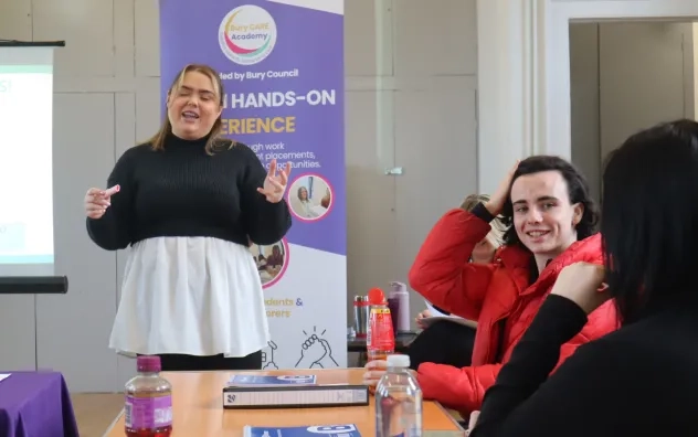 Bury Care Academy trainer stood at the front of the class room speaking while attendee is seen smiling while listening to another participant talking to the trainer